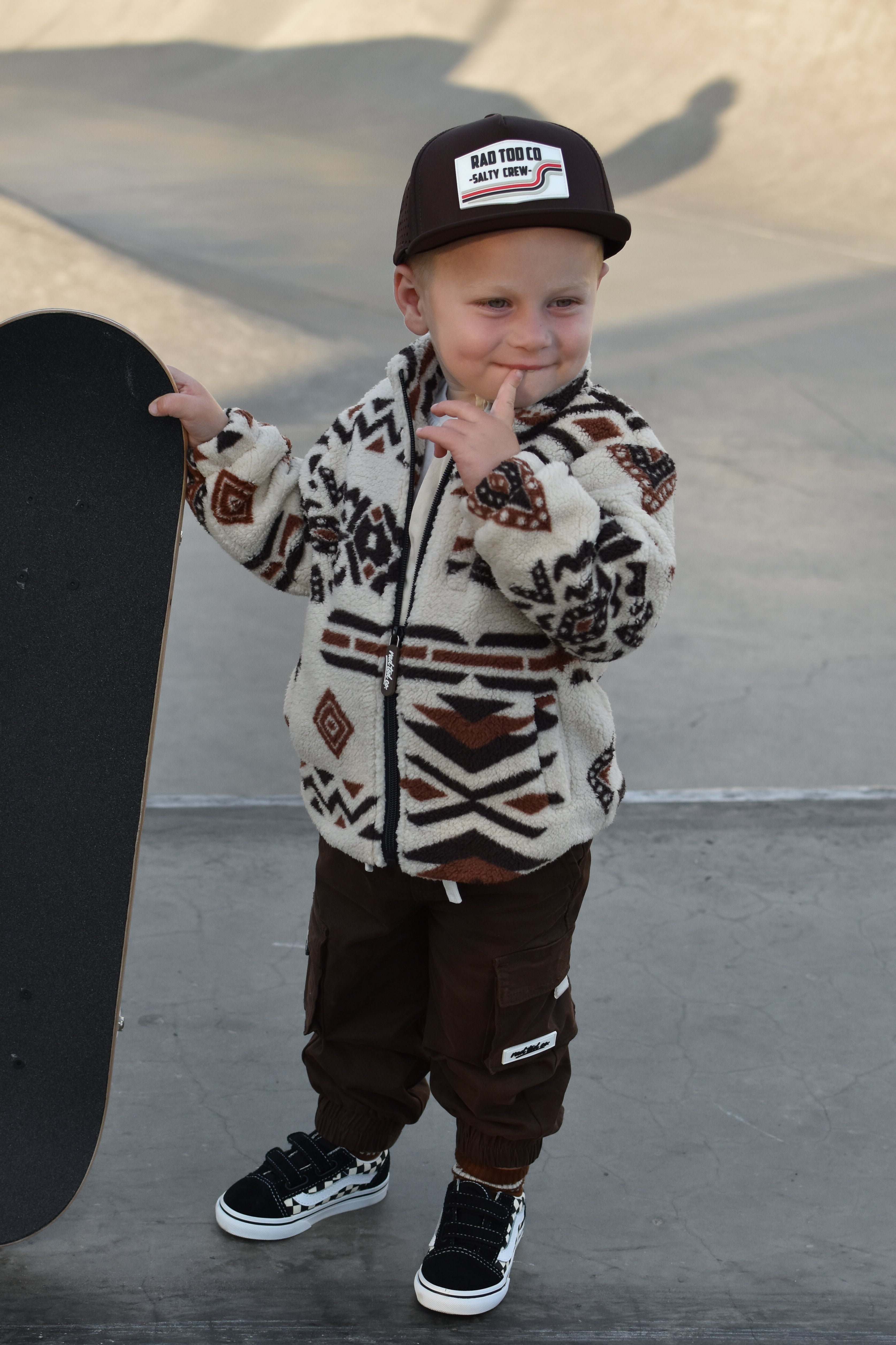 A toddler stands at a skatepark wearing a cream and brown patterned sherpa zip-up hoodie, dark brown pants, a black cap with a 'RAD TOD CO' patch, and black sneakers, holding a skateboard.