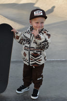 A toddler stands at a skatepark wearing a cream and brown patterned sherpa zip-up hoodie, dark brown pants, a black cap with a 'RAD TOD CO' patch, and black sneakers, holding a skateboard.