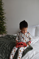 A young boy sits on a bed wearing white pajamas with a milk cookies and chaos print, holding a red toy car. A dark green textured blanket is draped over the bed, and a decorated Christmas tree is visible in the background against a gray wall.
