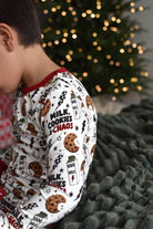 A child wearing a white long-sleeve pajama top with a red collar and a 'Milk, Cookies & Chaos' holiday print, sitting on a dark textured blanket in front of a blurred Christmas tree with warm lights.