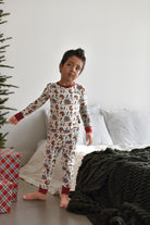 A young boy wearing a long-sleeve Christmas-themed pajama set with a brown and red milk cookies and chaos print by Rad TOddler on a white background, standing in a bedroom next to a bed and a Christmas tree.