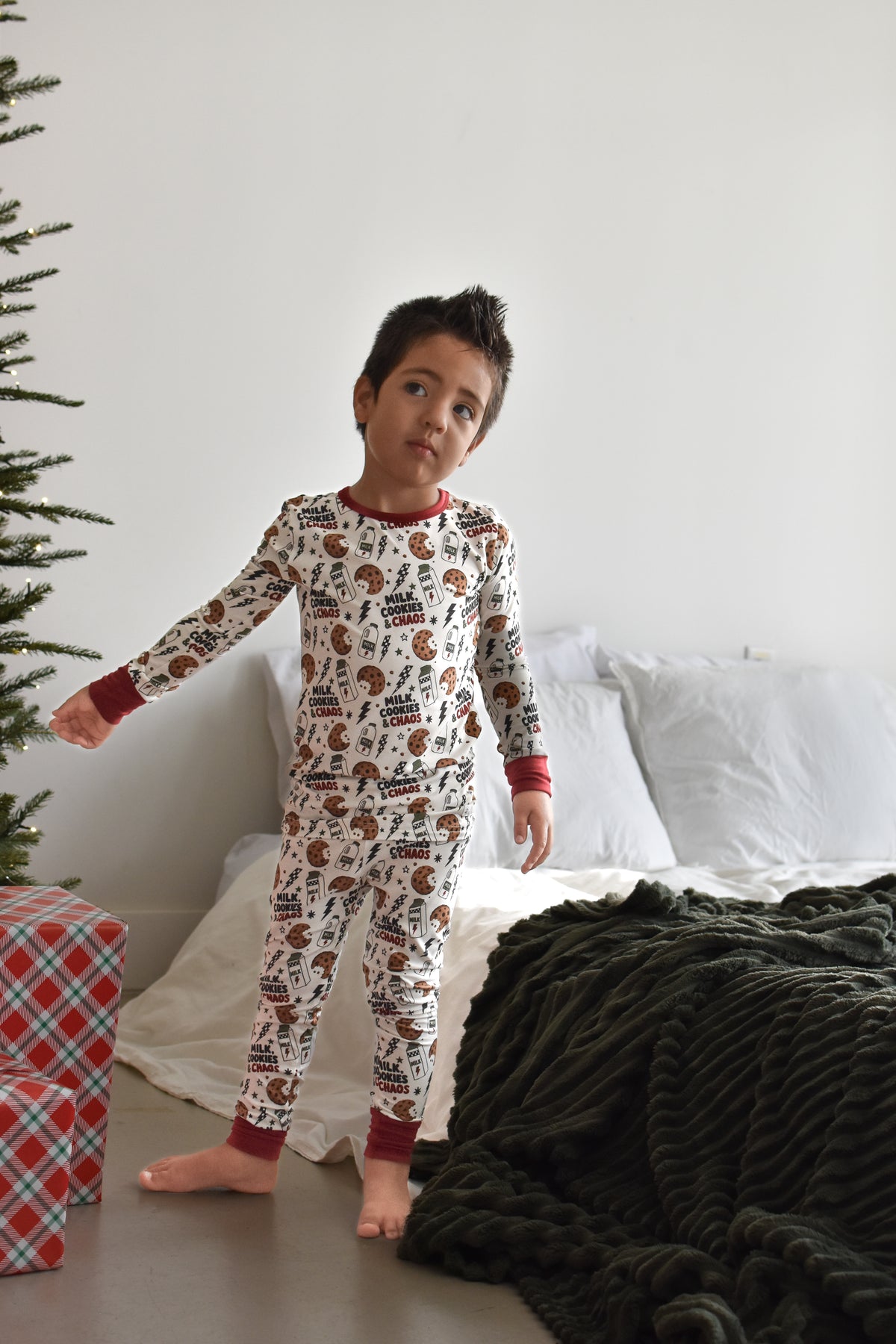 A young boy wearing a long-sleeve Christmas-themed pajama set with a brown and red milk cookies and chaos print by Rad TOddler on a white background, standing in a bedroom next to a bed and a Christmas tree.