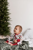 A baby sitting on a textured green blanket in front of a decorated Christmas tree, wearing a white and red holiday print one-piece pajama.