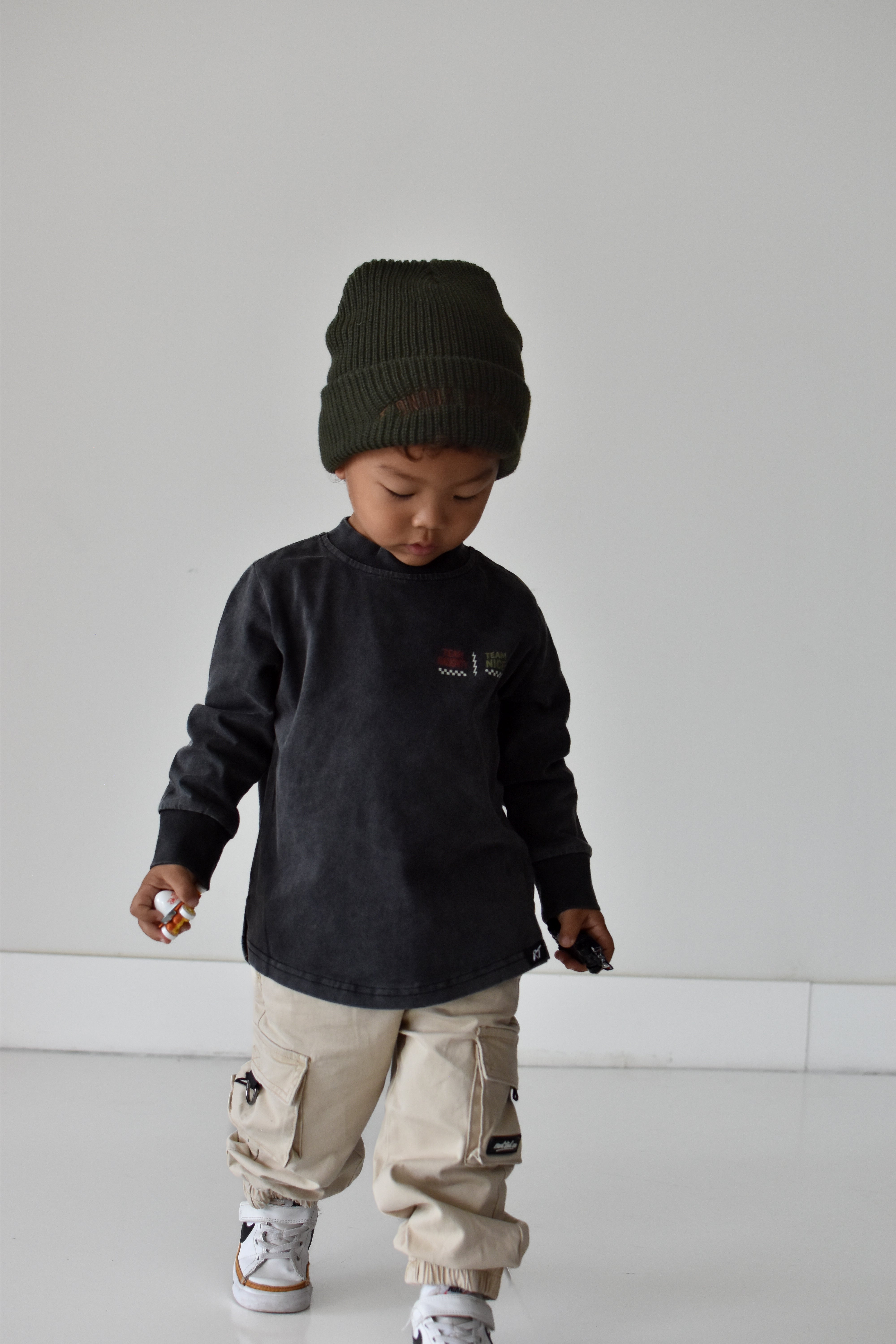 A toddler wearing a dark charcoal long-sleeve shirt with a small chest graphic, beige cargo pants, and a dark olive green knit beanie, standing against a minimalist light gray background.
