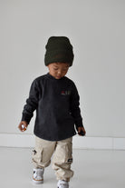 A toddler wearing a dark charcoal long-sleeve shirt with a small chest graphic, beige cargo pants, and a dark olive green knit beanie, standing against a minimalist light gray background.