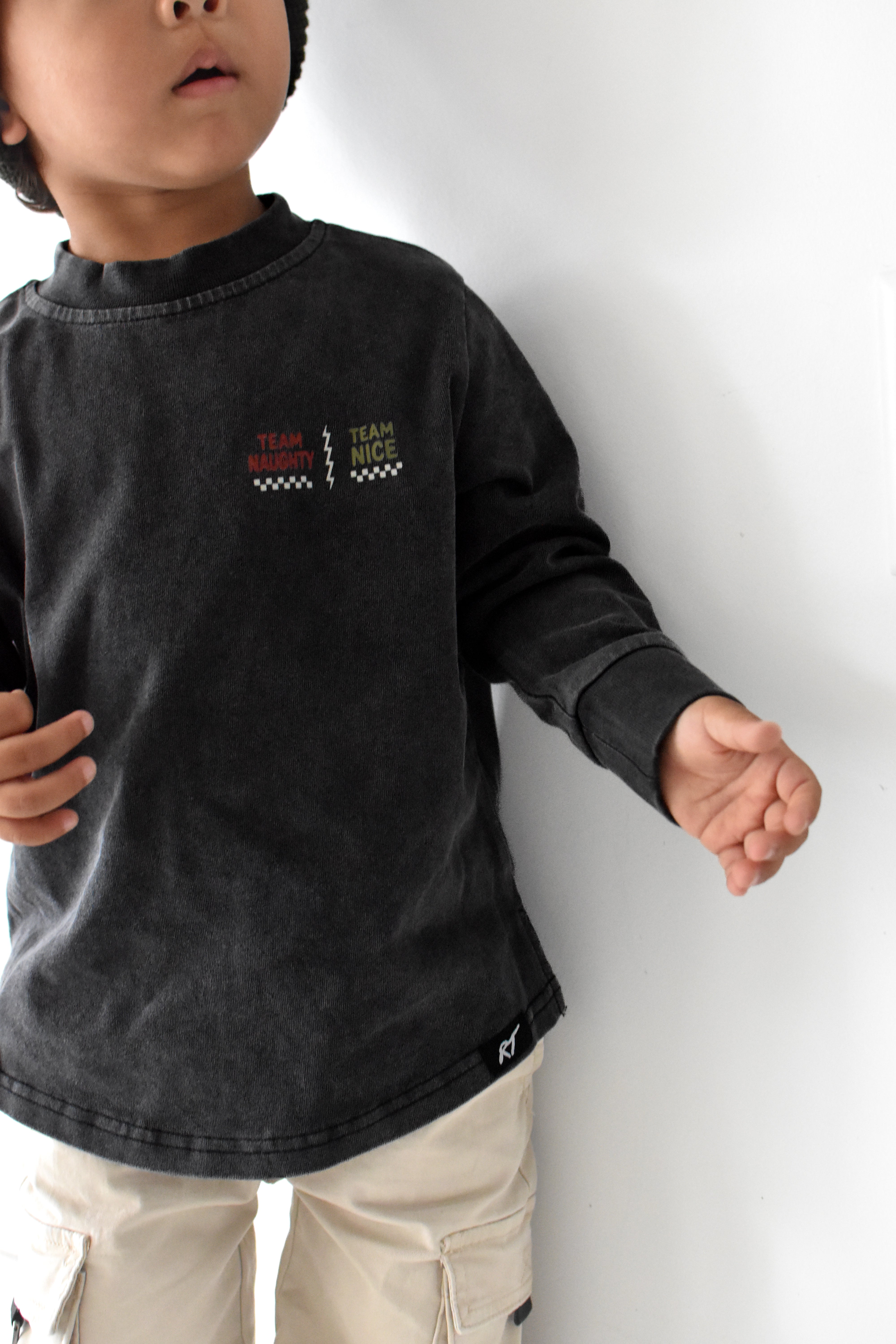 A child wearing a dark charcoal gray long-sleeve t-shirt with a small graphic on the left chest, paired with beige cargo pants, standing against a plain white wall.