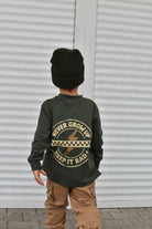 A child viewed from behind wearing an olive green long-sleeve shirt with a tan graphic print on the back, paired with a black knit beanie and tan cargo pants, standing in front of a white corrugated metal wall.