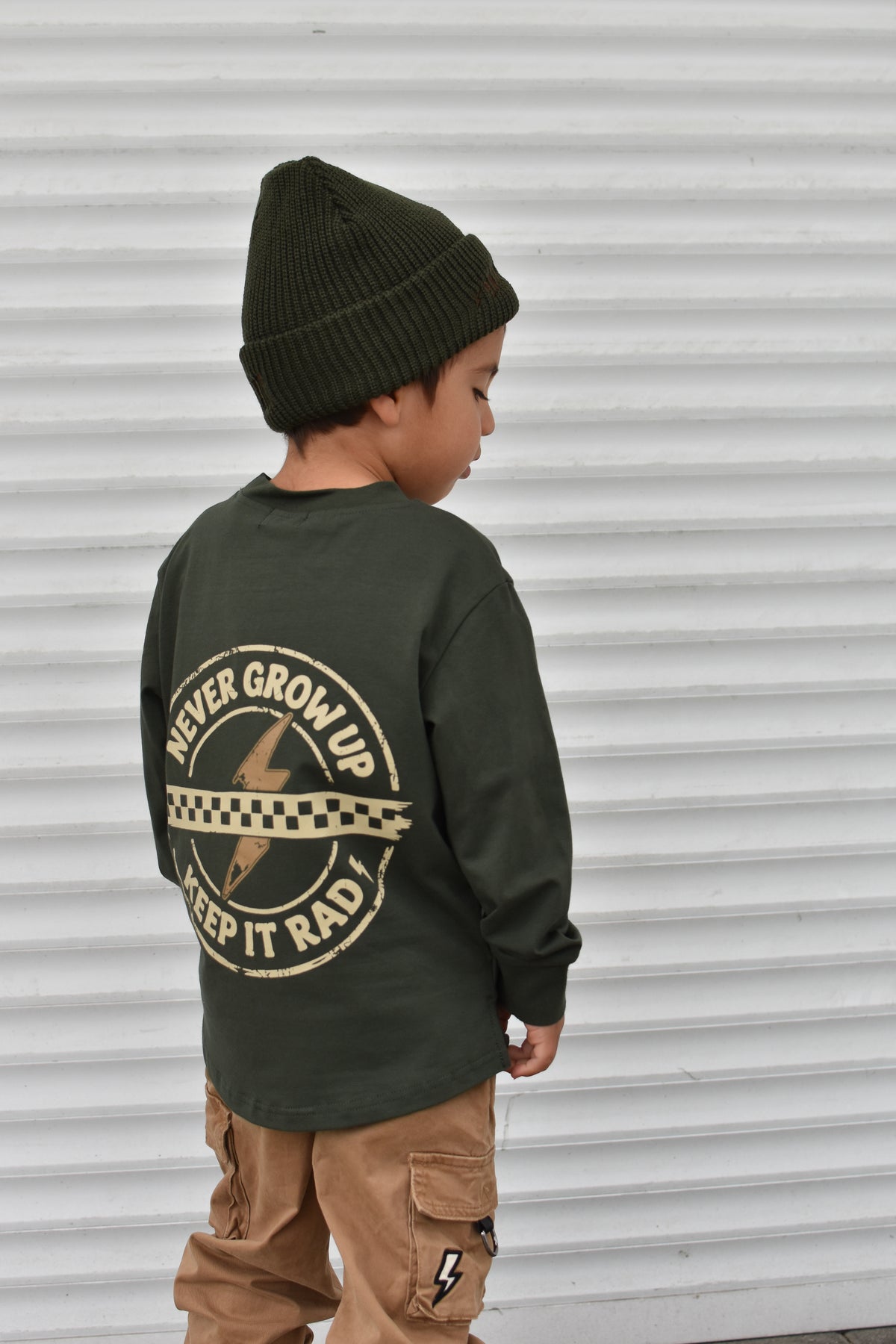 A child wearing an olive green long-sleeve shirt with a large graphic print on the back, paired with a dark green beanie and tan cargo pants, standing in front of a white corrugated metal wall.