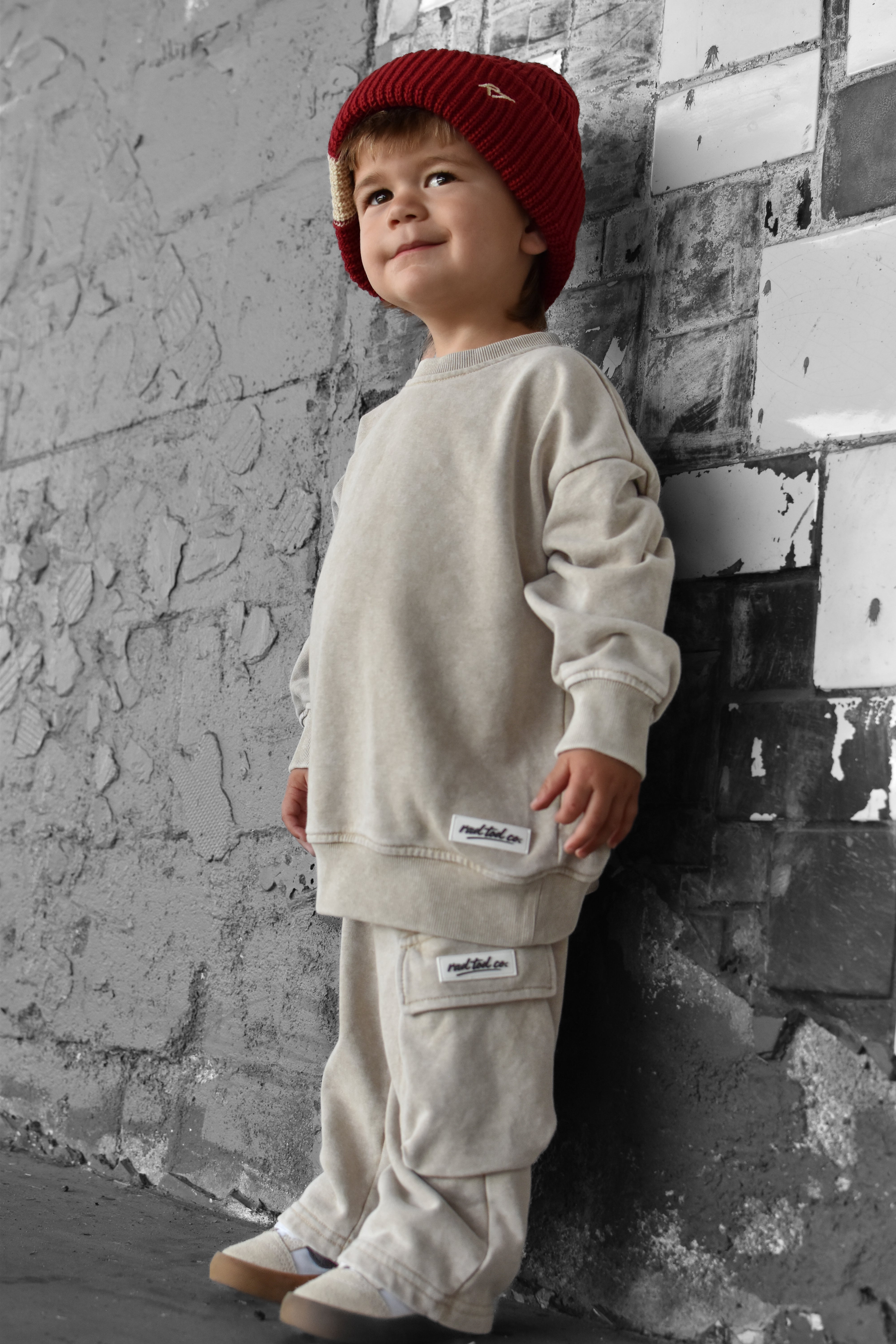 A toddler stands against a textured gray brick wall wearing a cream-colored sweatshirt and matching cargo pants, paired with a red knit beanie.