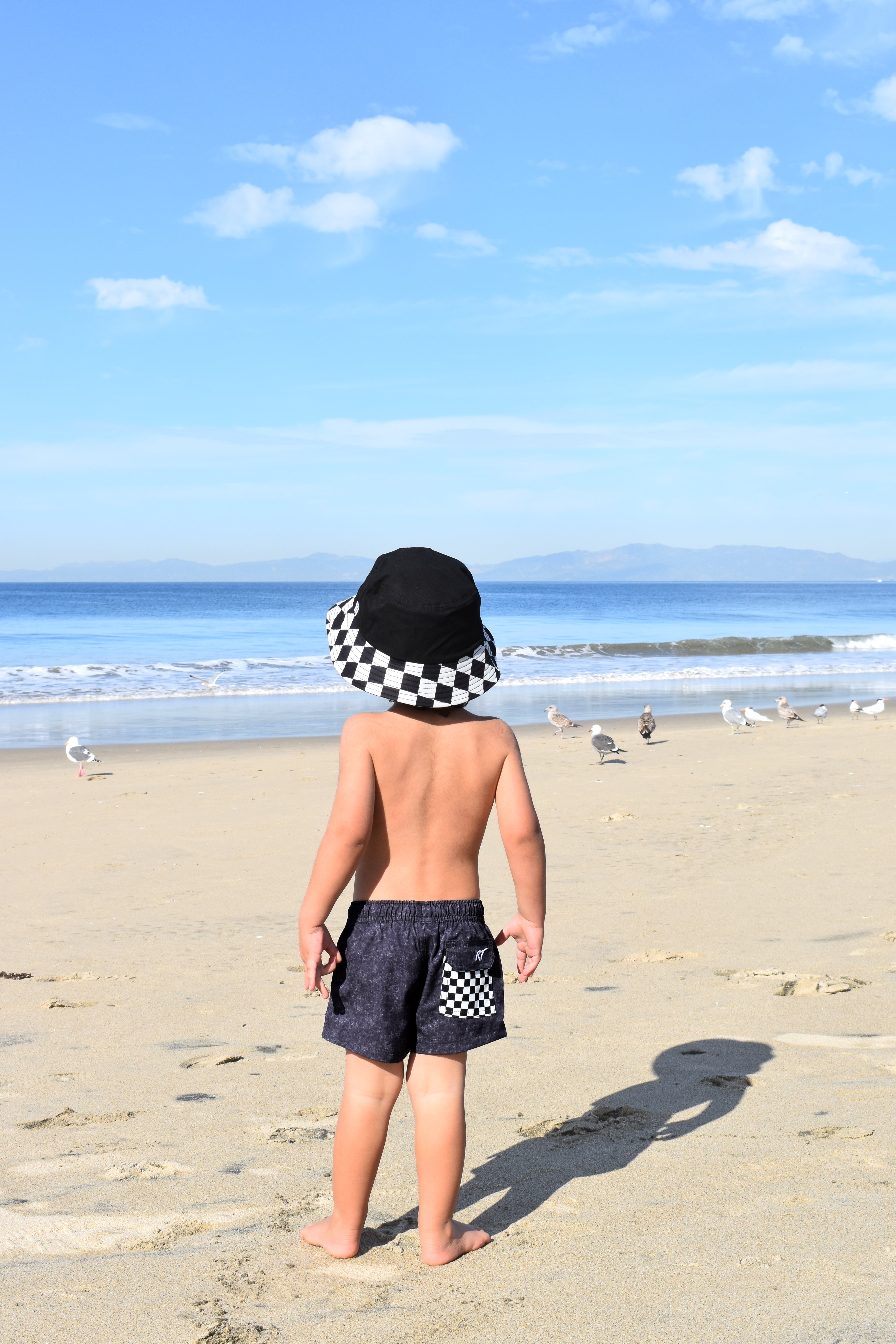 Child standing on a beach wearing a checkered hat and shorts, looking at the ocean.