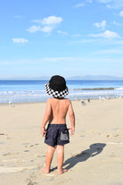 Child standing on a beach wearing a checkered hat and shorts, looking at the ocean.