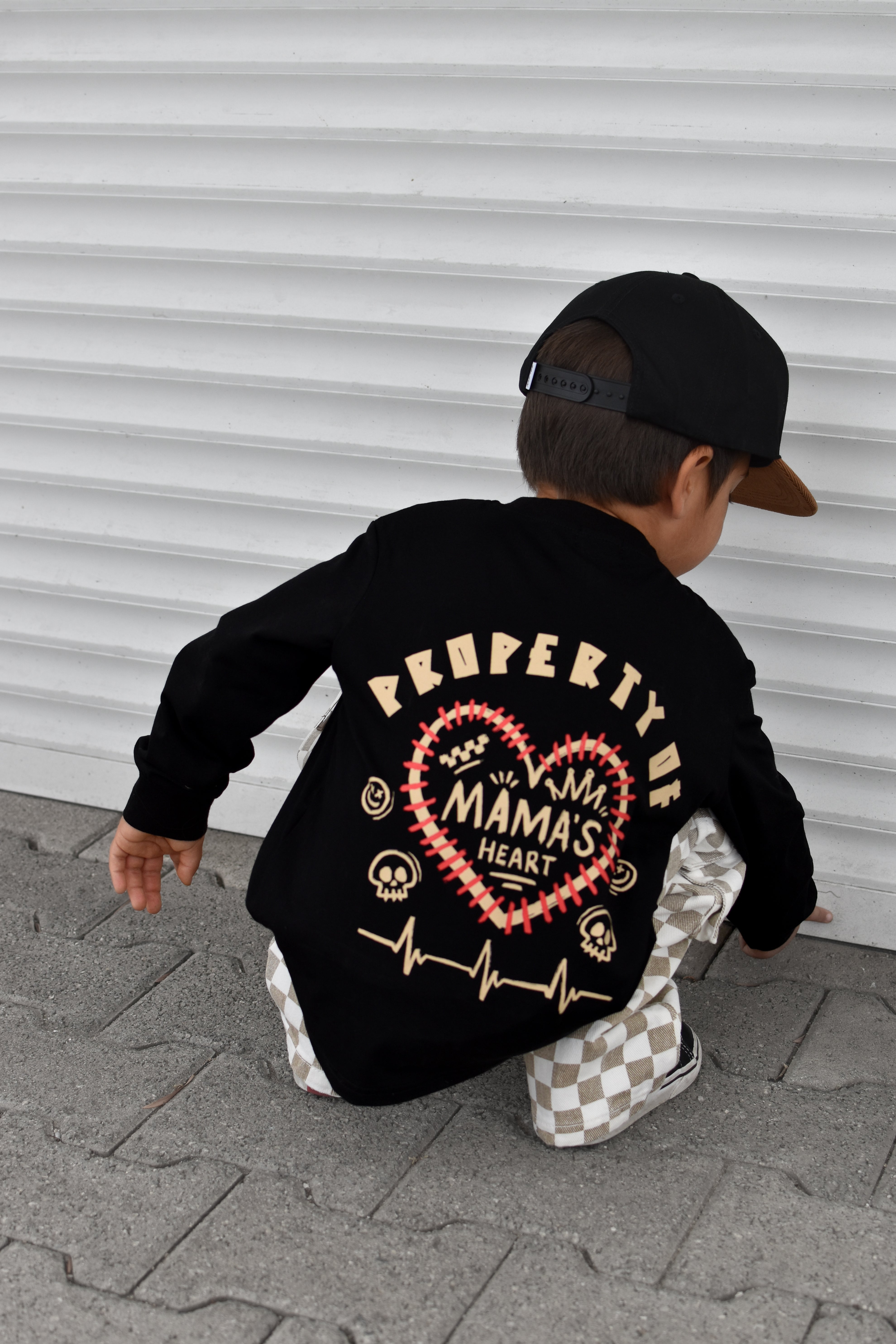 A toddler wearing a black long-sleeve shirt with a large graphic on the back, paired with a black cap and checkered pants, crouching in front of a white corrugated metal wall.
