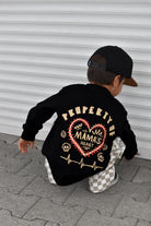 A toddler wearing a black long-sleeve shirt with a large graphic on the back, paired with a black cap and checkered pants, crouching in front of a white corrugated metal wall.