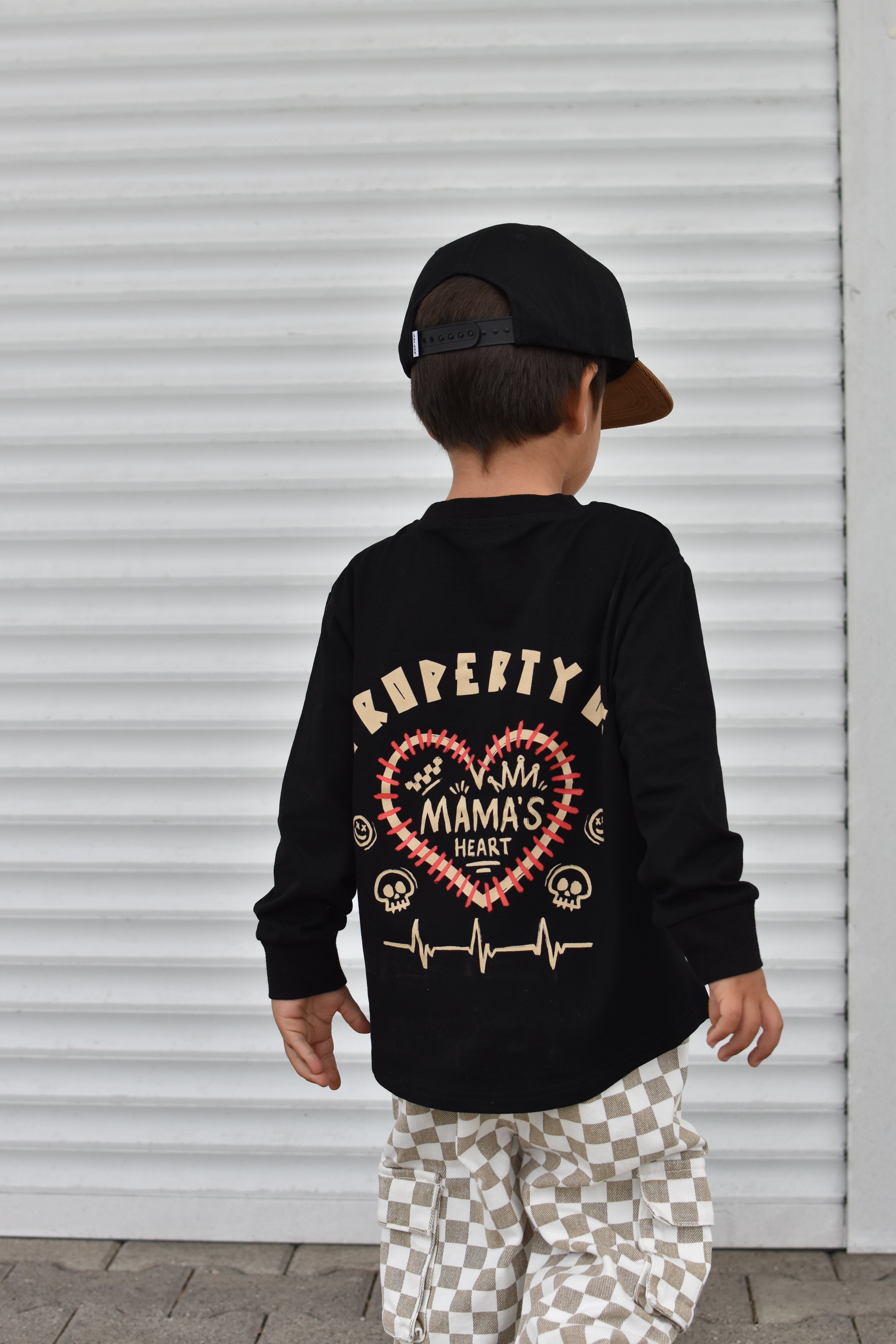 A child wearing a black long-sleeve shirt with a large red and gold heart graphic and the words 'PROPERTY OF MAMA'S HEART' on the back, paired with beige-and-white checkered pants and a black cap, standing in front of a white corrugated metal wall.