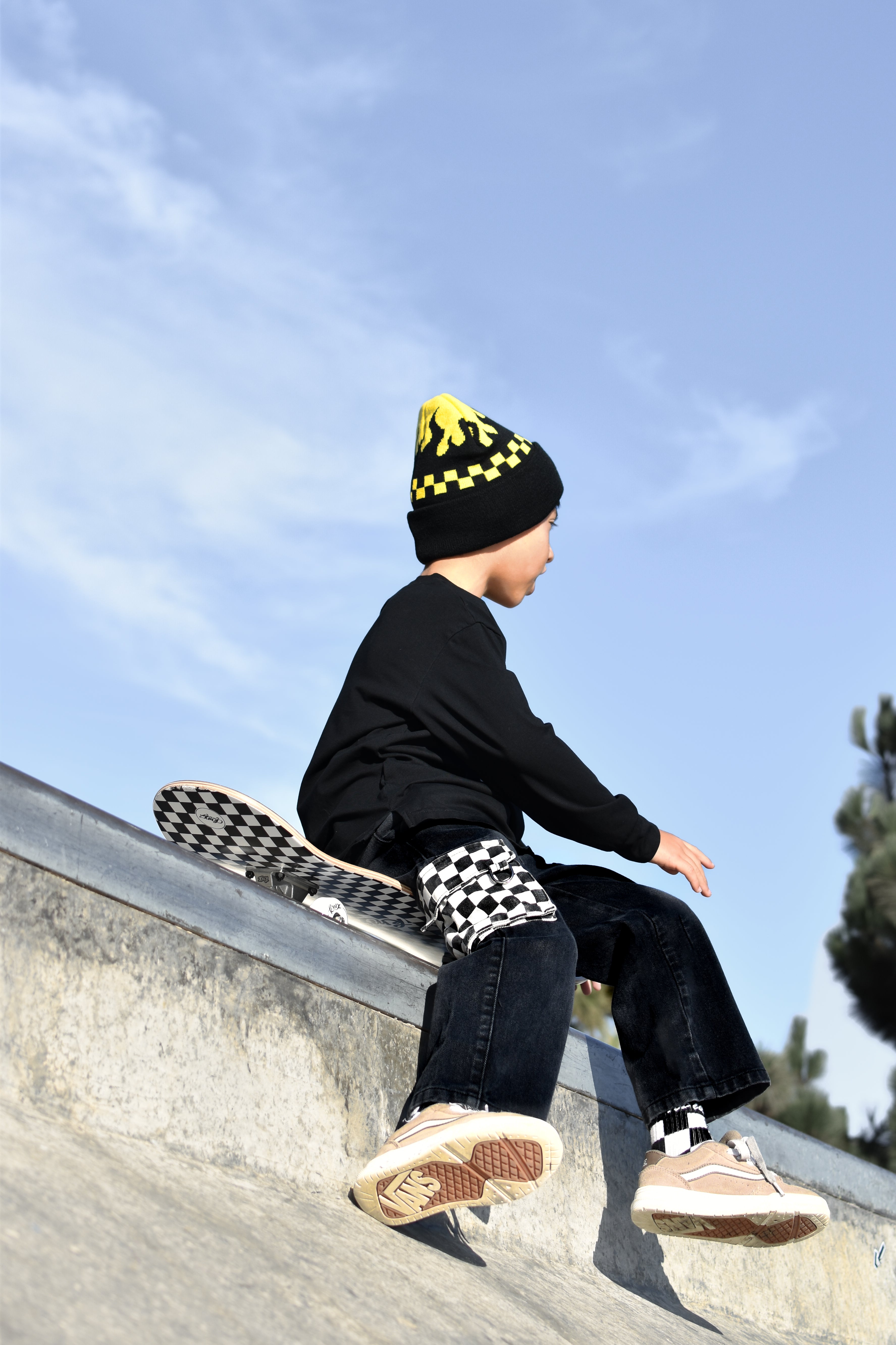 A child wearing a black beanie with yellow details, a black long-sleeve top, and dark jeans sits on the edge of a concrete skate ramp under a blue sky. The child is wearing Vans sneakers and has a Vans skateboard resting on the ramp behind them.