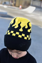 A child wearing a black knit beanie with a yellow flame pattern and a yellow-and-black checkered band, photographed outdoors with a blurred skatepark background.