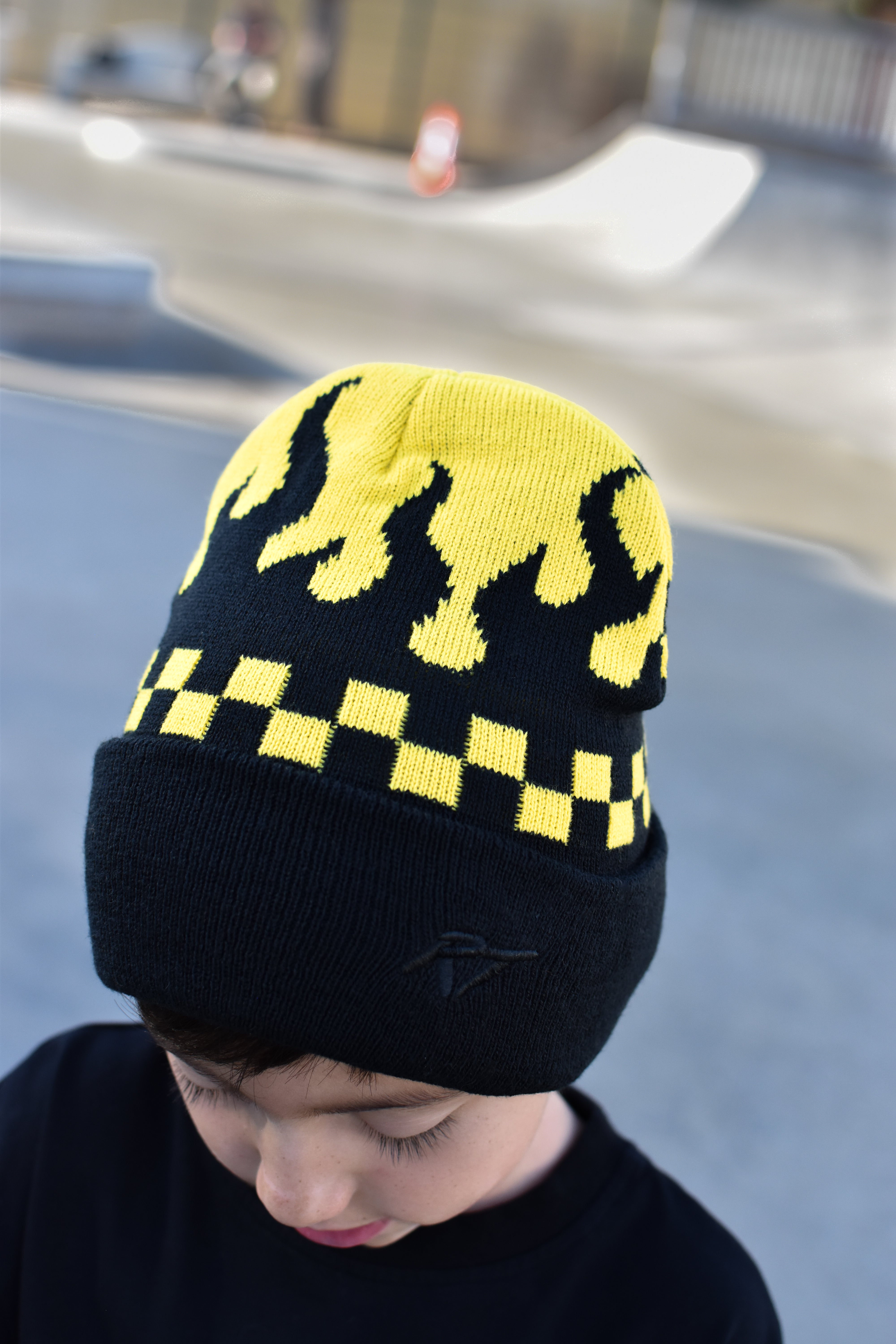 A child wearing a black knit beanie with a yellow flame pattern and a yellow-and-black checkered band, photographed outdoors with a blurred skatepark background.