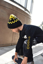 A child wearing a black beanie with yellow graphic details, a black long-sleeve shirt with yellow smiley face graphics on the sleeve, and black pants, crouching on a skateboard outdoors.