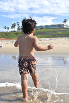 Child playing in shallow water at the beach while wearing Rad Toddler swimmies with palm trees and buildings in the background.