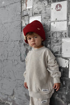 A young child wearing a cream-colored sweatshirt and matching pants with a red knit beanie, standing against a textured gray brick wall.