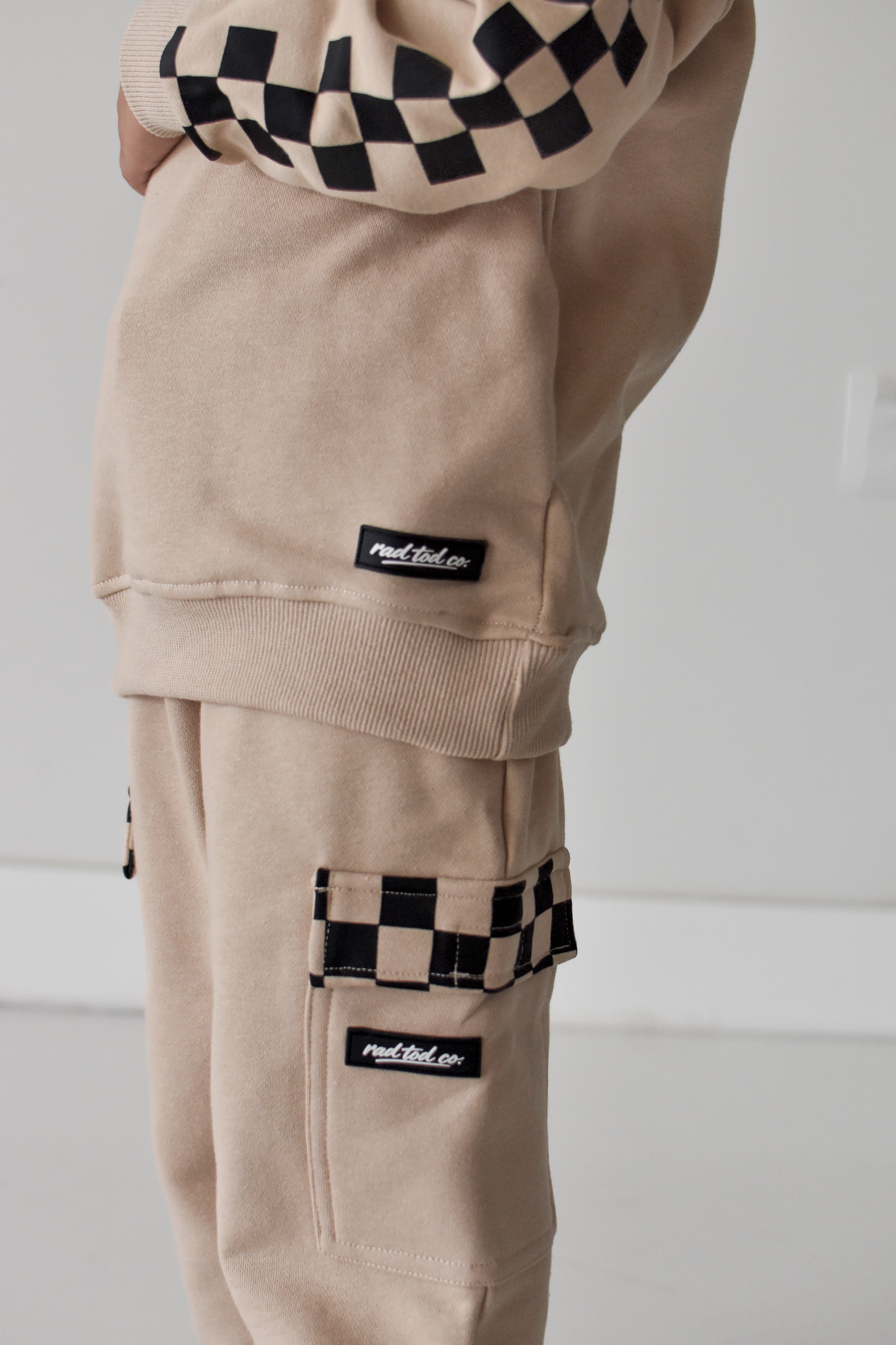 A close-up of a person wearing beige cargo sweatpants with black-and-white checkerboard accents on the pocket trim and cuffs, plus matching beige sweatshirt shown at the top.
