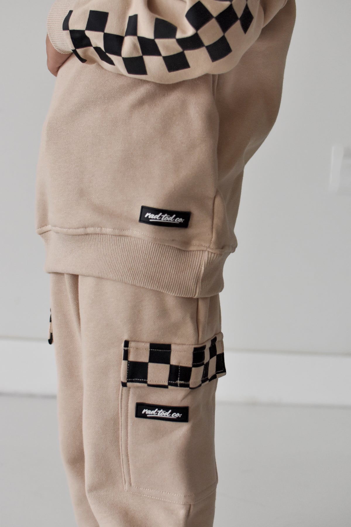 A close-up of a person wearing beige cargo sweatpants with black-and-white checkerboard accents on the pocket trim and cuffs, plus matching beige sweatshirt shown at the top.