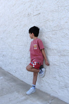 Child wearing a red t-shirt and checkered shorts against a white brick wall.