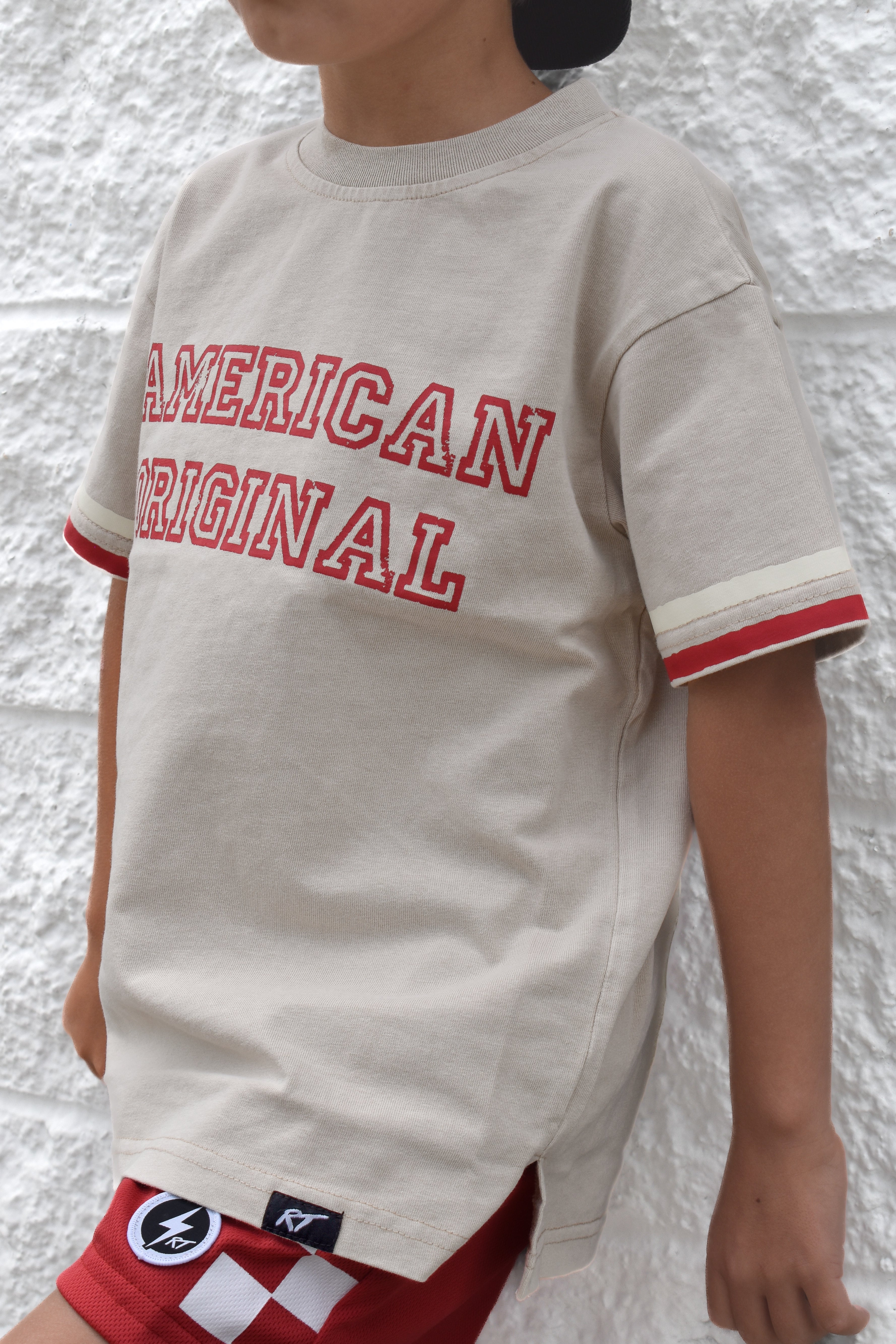 Child wearing a beige t-shirt with 'American Original' text against a textured white wall.
