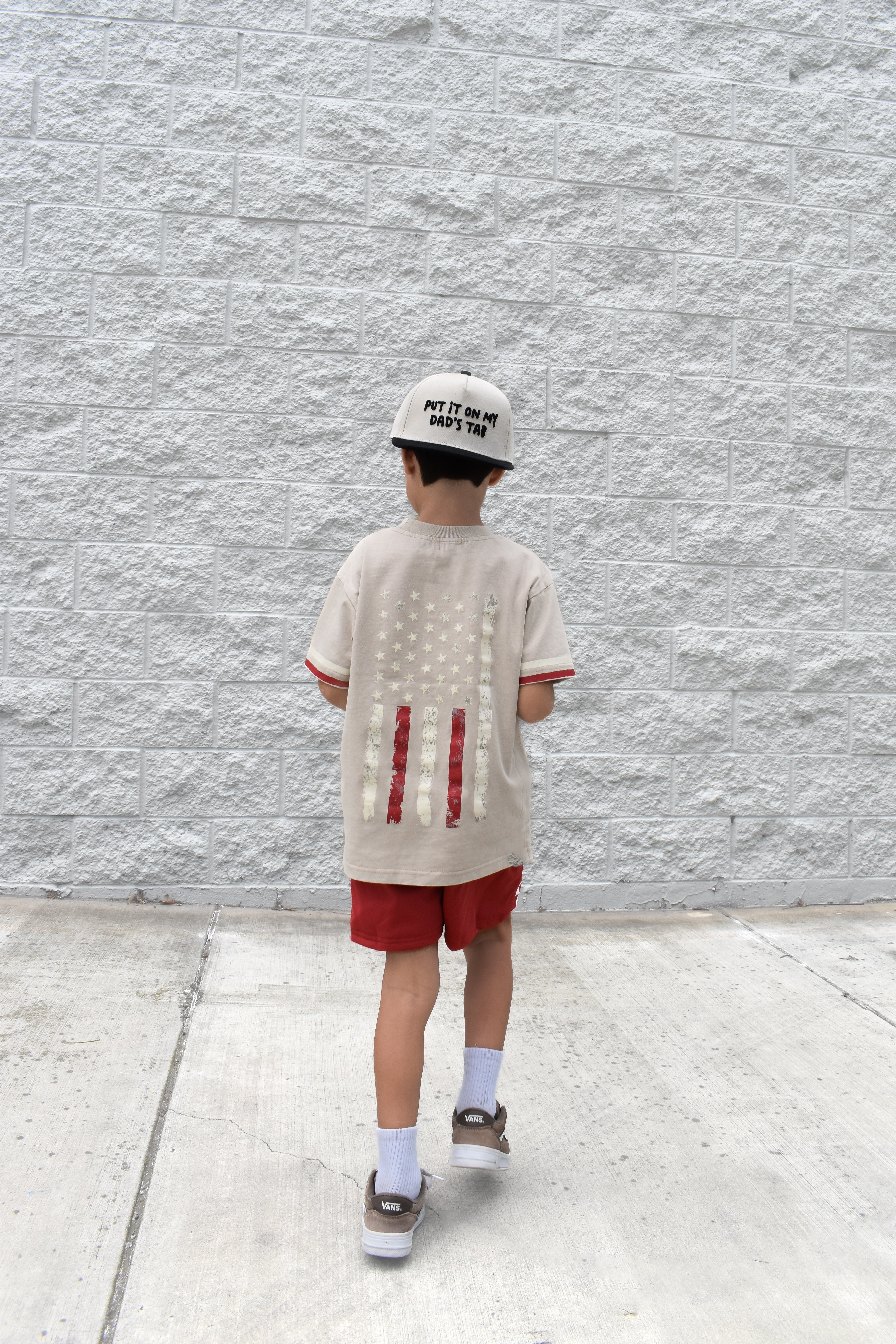 Child wearing a beige t-shirt with red designs and red shorts against a gray textured wall.