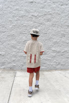 Child wearing a beige t-shirt with red designs and red shorts against a gray textured wall.