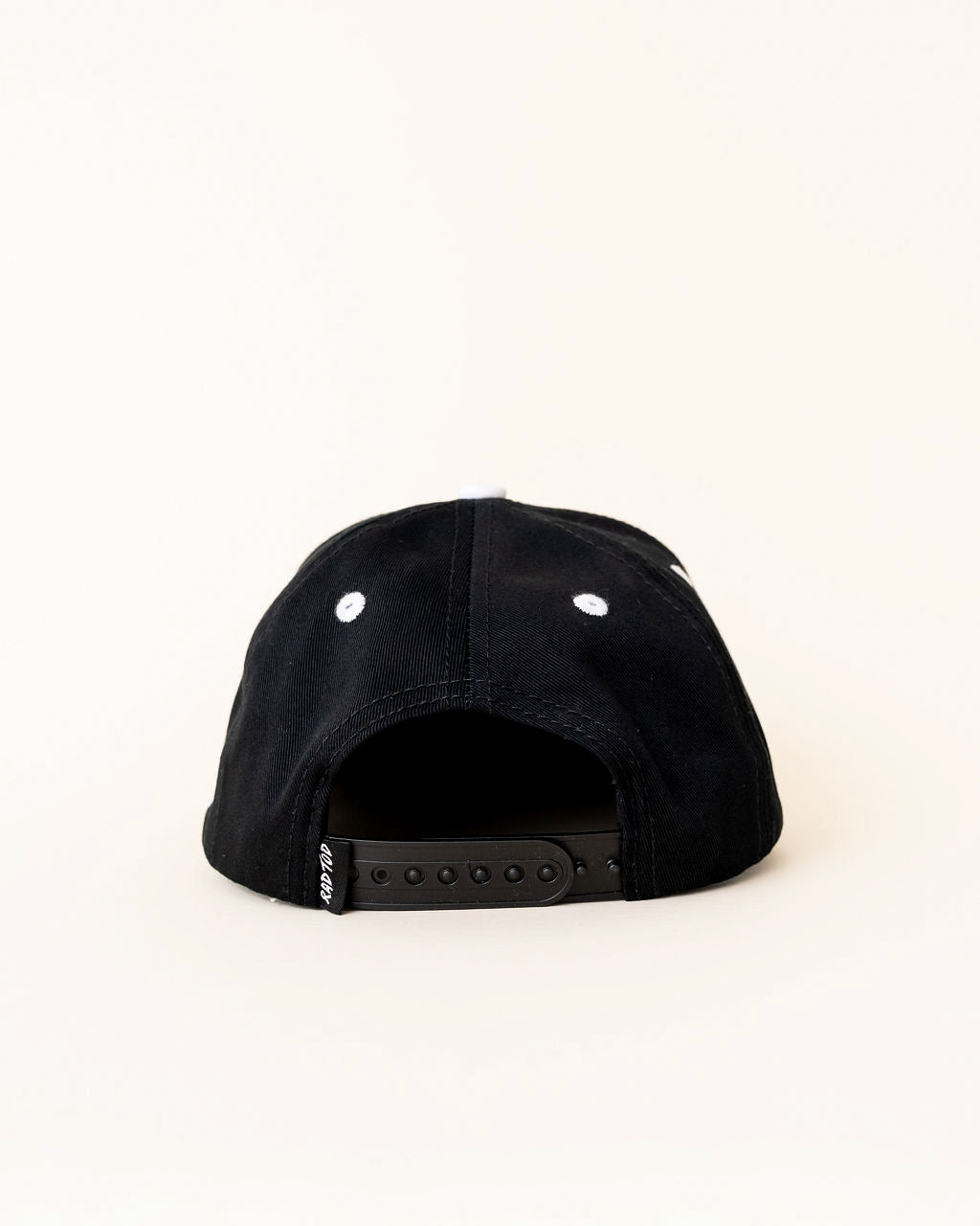 A black snapback cap shown from the back, featuring white embroidered eyelets and a black adjustable strap with a small white brand tag.