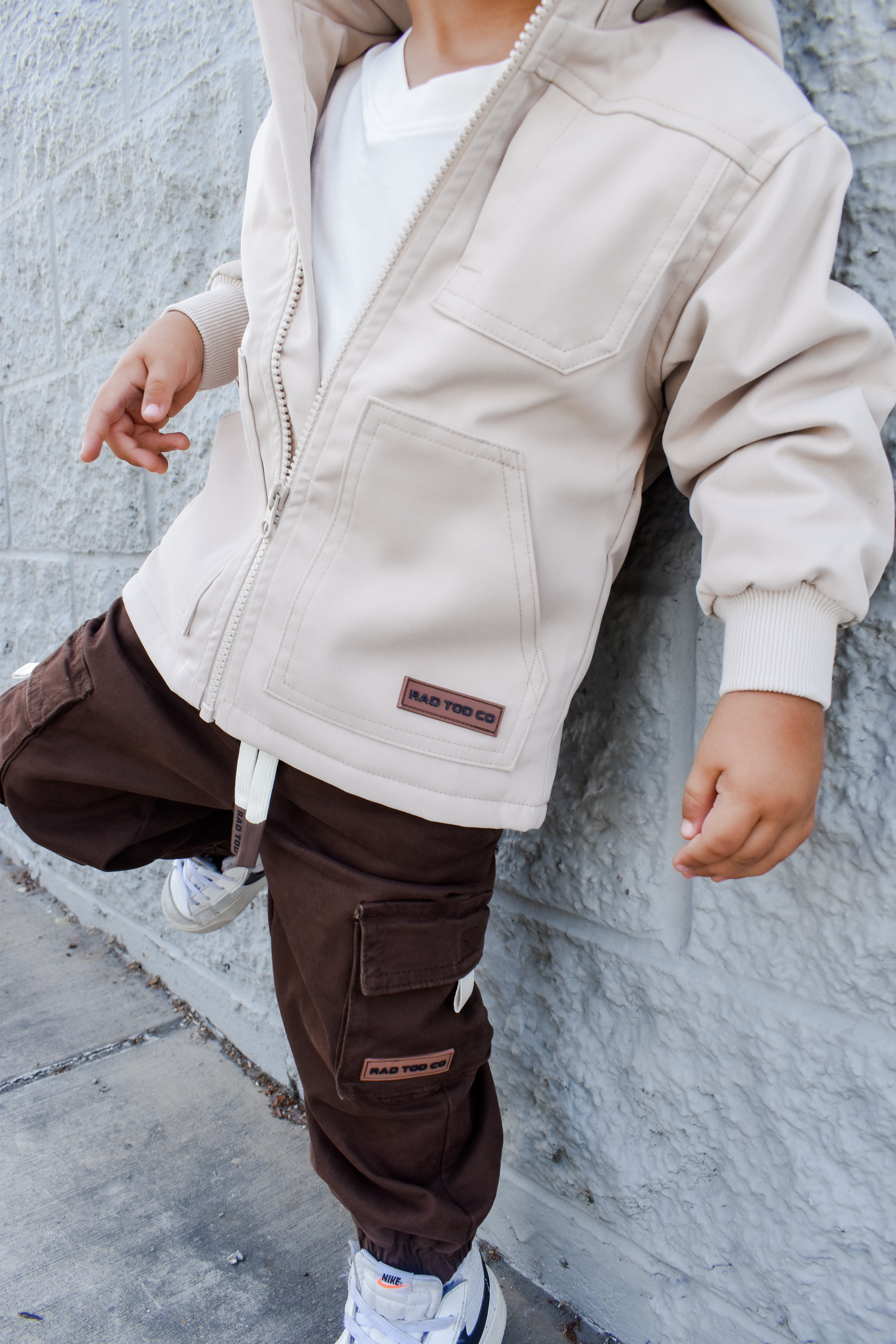 Child wearing a beige jacket and brown pants against a gray wall.