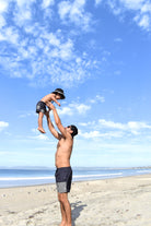 Man lifting a child in the air on a beach with a clear blue sky while sporting some awesome rad toddler gear