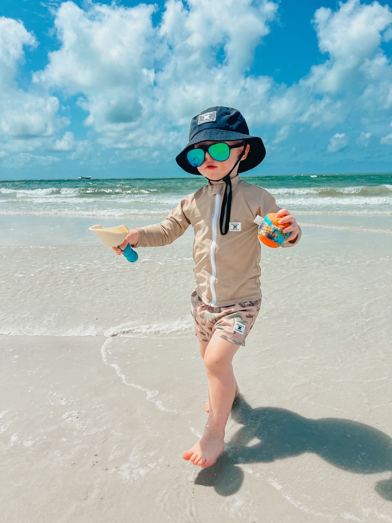 beach day wearing Rad Toddlers 'Sand' Rashguard Shirt with front zipper and UPF 50+ protection, perfect for pairing with board shorts for water activities.