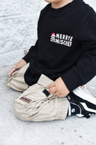 Child wearing a black sweatshirt with 'Merry Mischief' text and beige pants on a light gray background