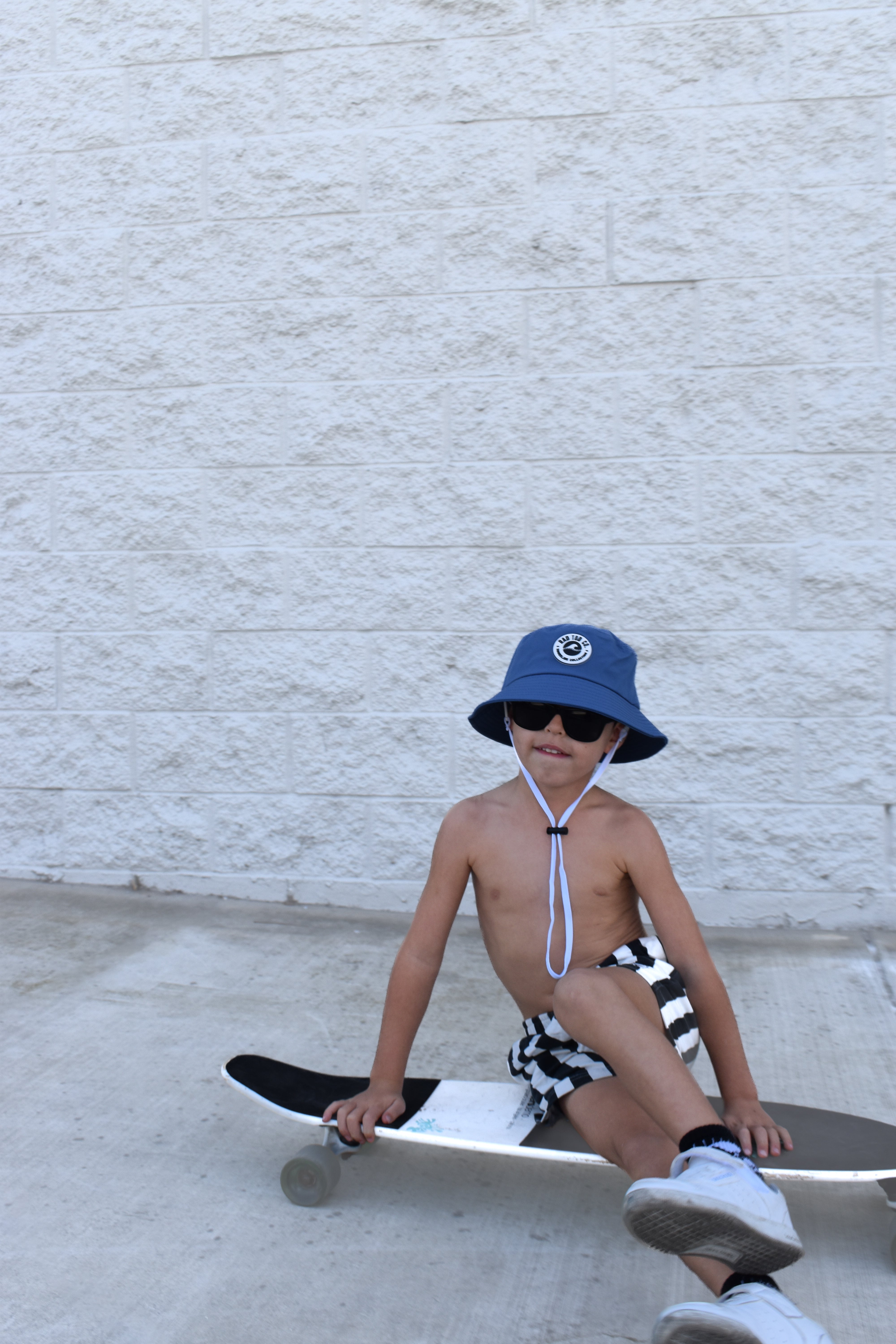 Child sitting on a skateboard wearing a blue bucket hat and sunglasses against a light gray wall.