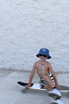 Child sitting on a skateboard wearing a blue bucket hat and sunglasses against a light gray wall.