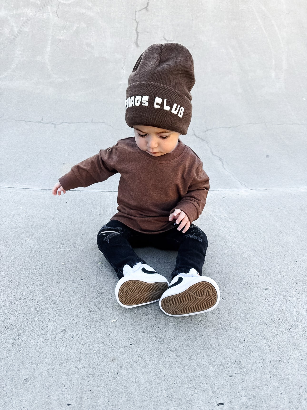 Child wearing a brown outfit with a beanie sitting on a concrete surface