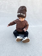 Child wearing a brown outfit with a beanie sitting on a concrete surface