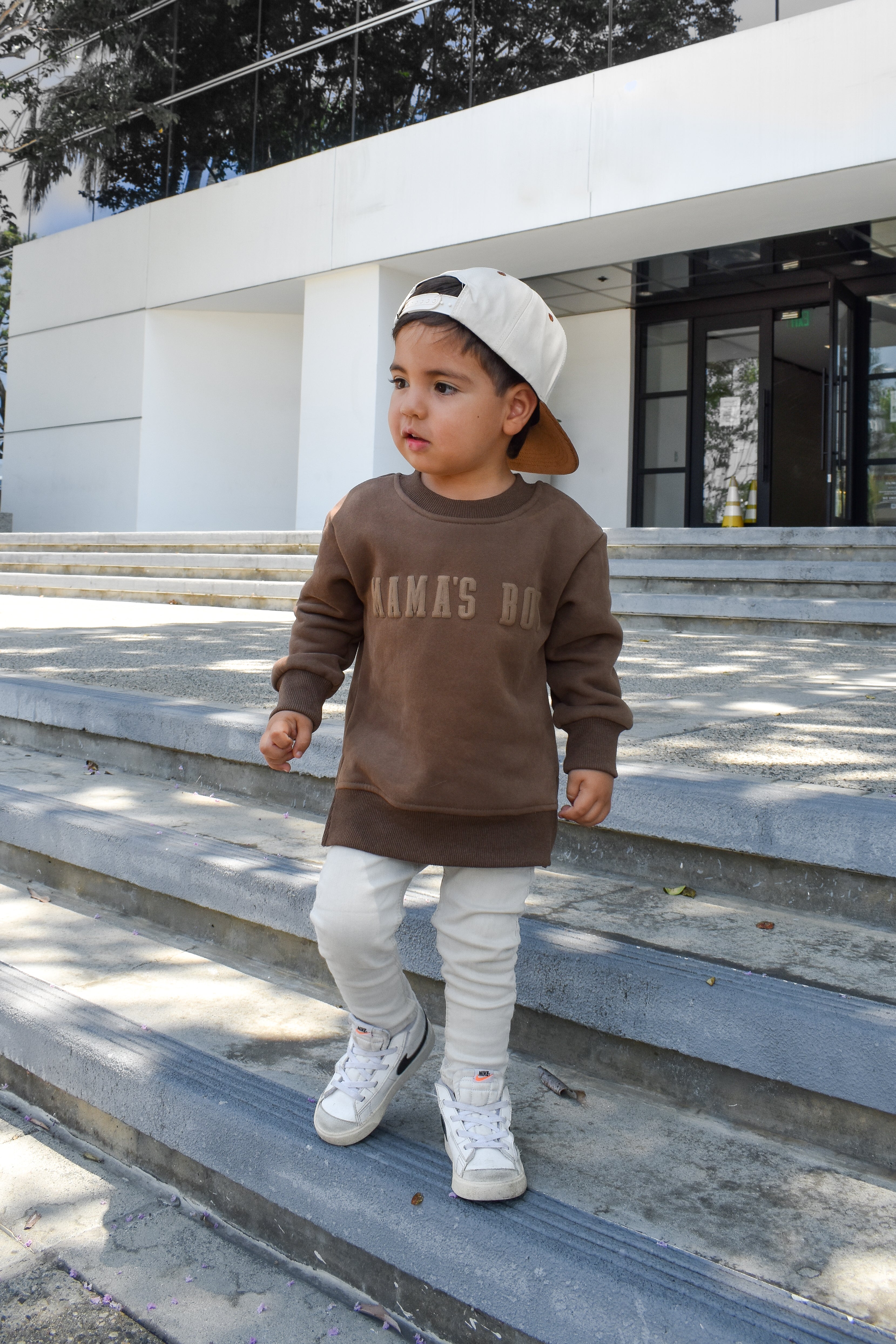 Child wearing brown “Mama’s Boy” crewneck with lighter brown lettering, relaxed fit, paired with casual bottoms.