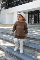 Child wearing brown “Mama’s Boy” crewneck with lighter brown lettering, relaxed fit, paired with casual bottoms.