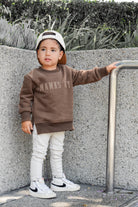 Child wearing brown “Mama’s Boy” crewneck with lighter brown lettering, relaxed fit, paired with casual bottoms.