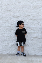 Child wearing a black t-shirt, striped shorts, and cap standing against a light stone wall.