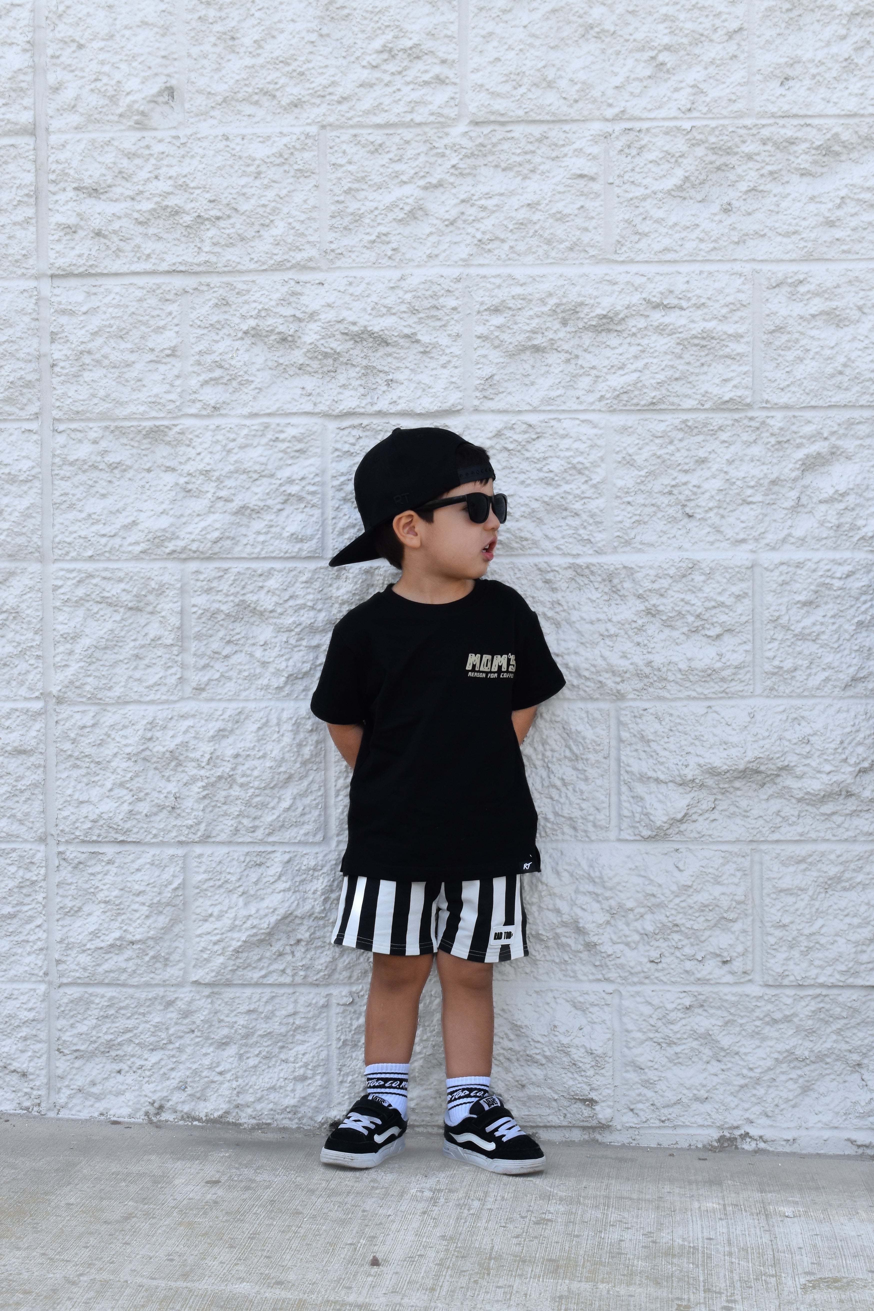 Child wearing a black t-shirt, striped shorts, and cap standing against a light stone wall.