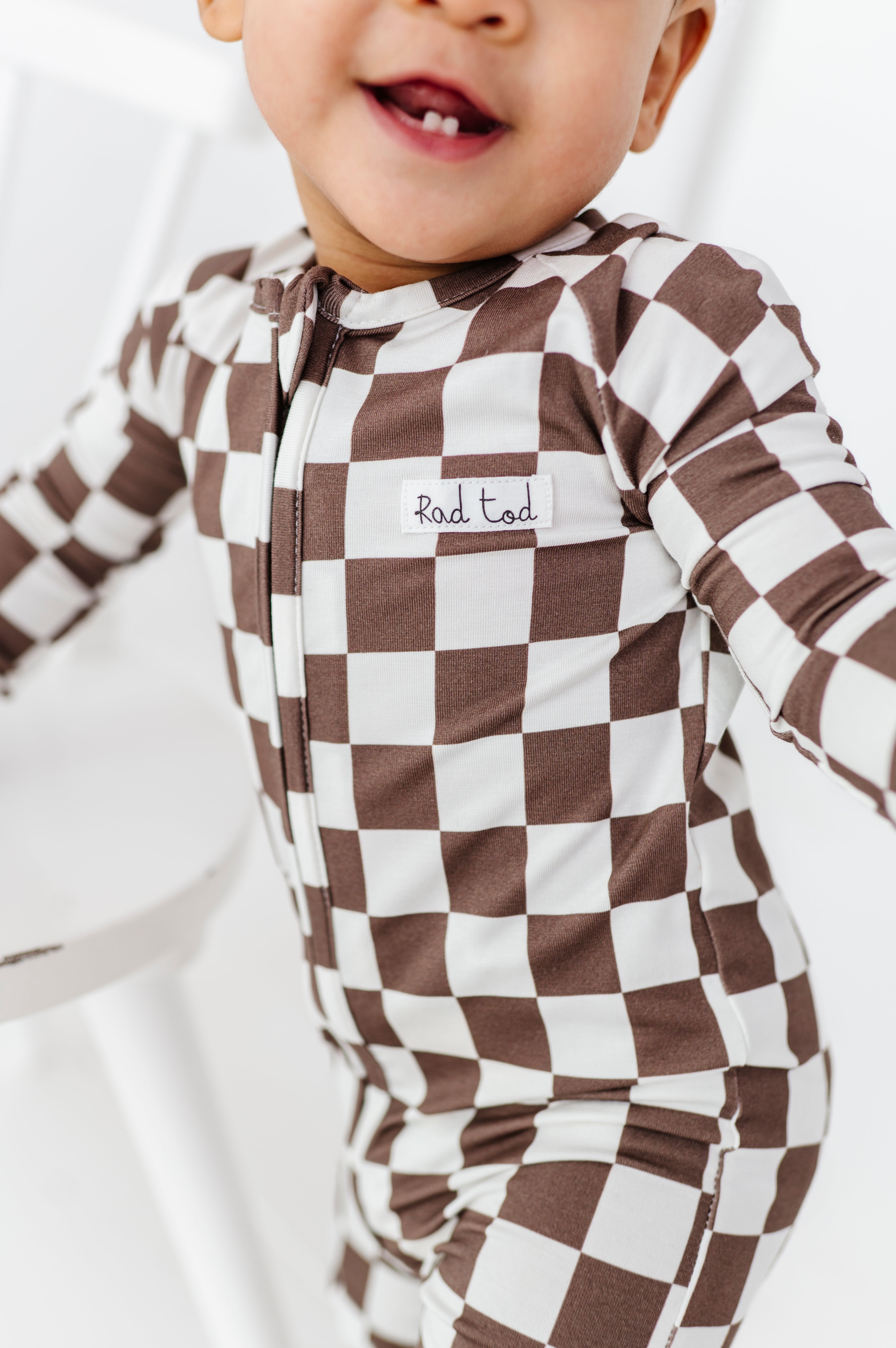 boy smiling wearing Bronze and white checkered bamboo pajamas for toddlers, featuring a two-way zipper and foldover footies by Rad Toddler.