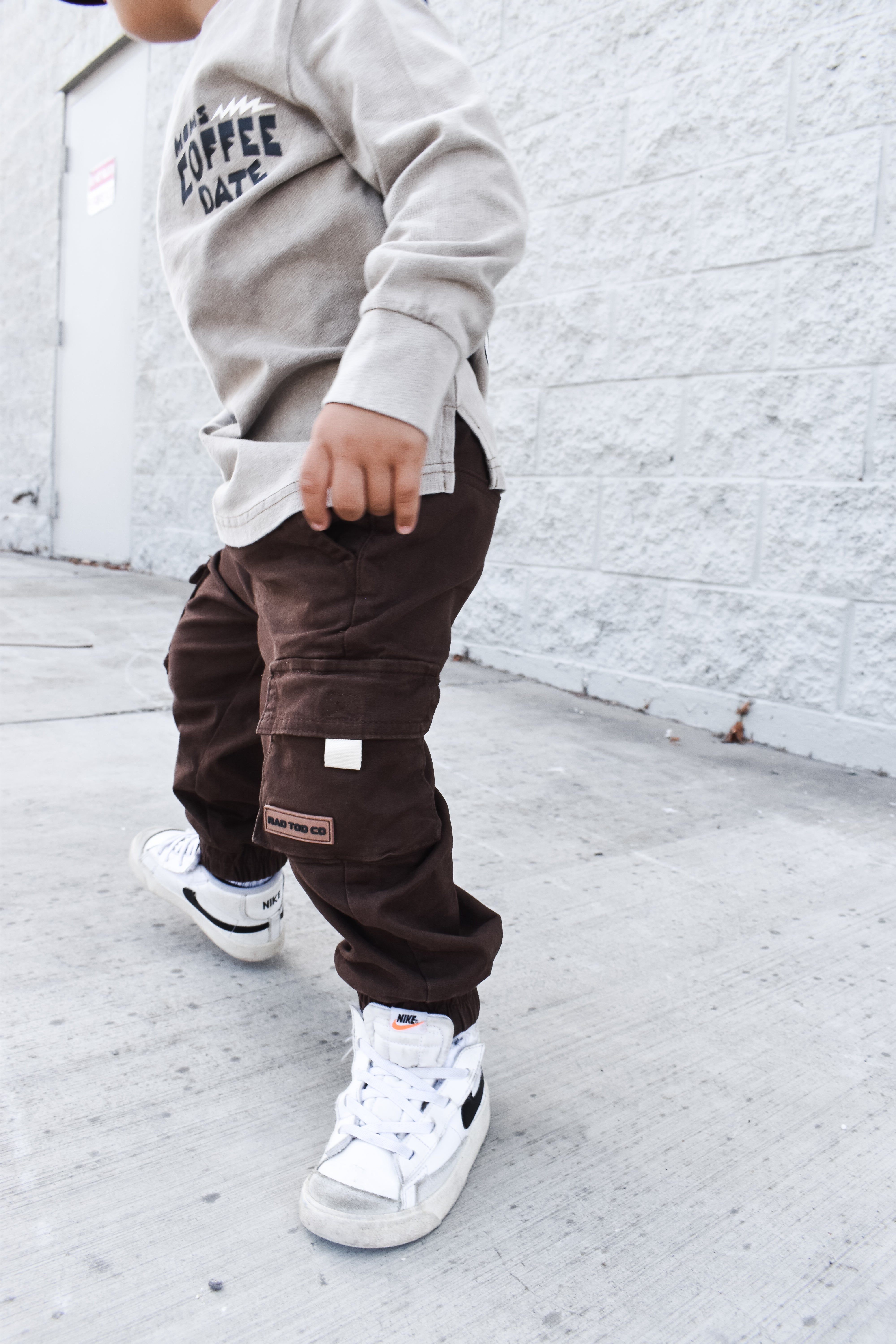 Child wearing a gray sweatshirt and brown cargo pants with white sneakers against a white brick wall by RAD TODDLER