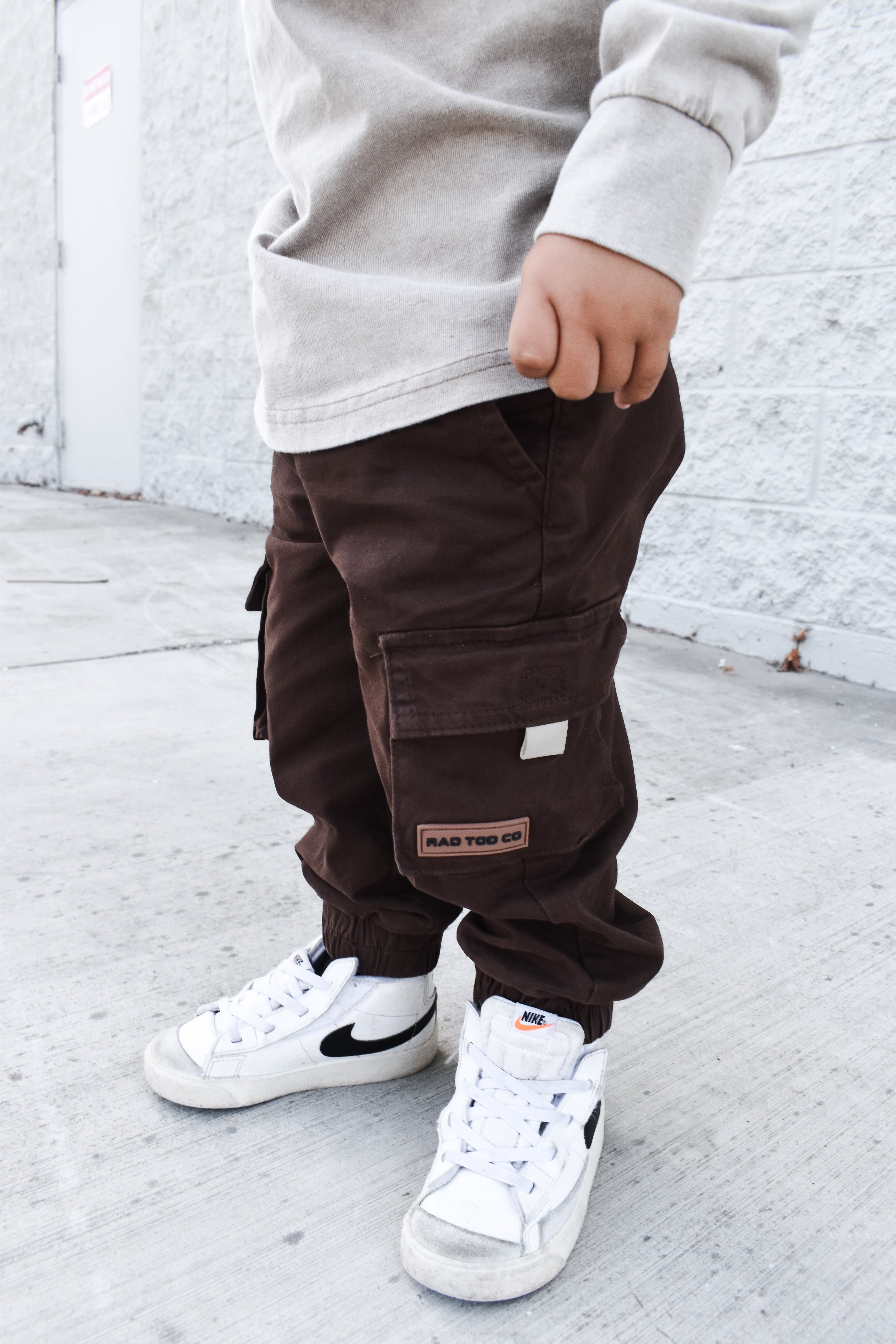 Child wearing brown cargo pants and white sneakers against a white brick wall.