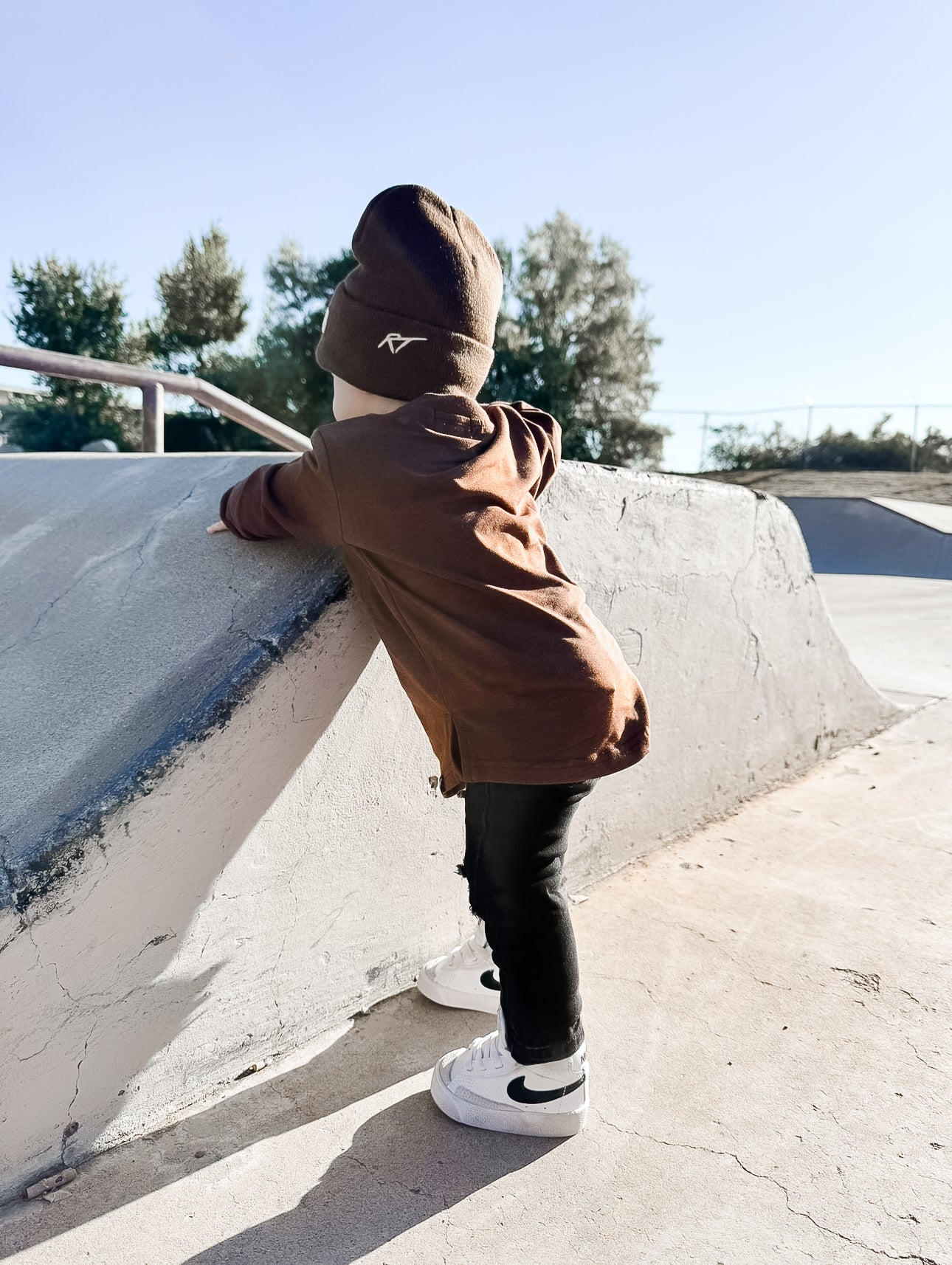 Child in a brown tee and black pants standing on a concrete ledge with trees in the background.