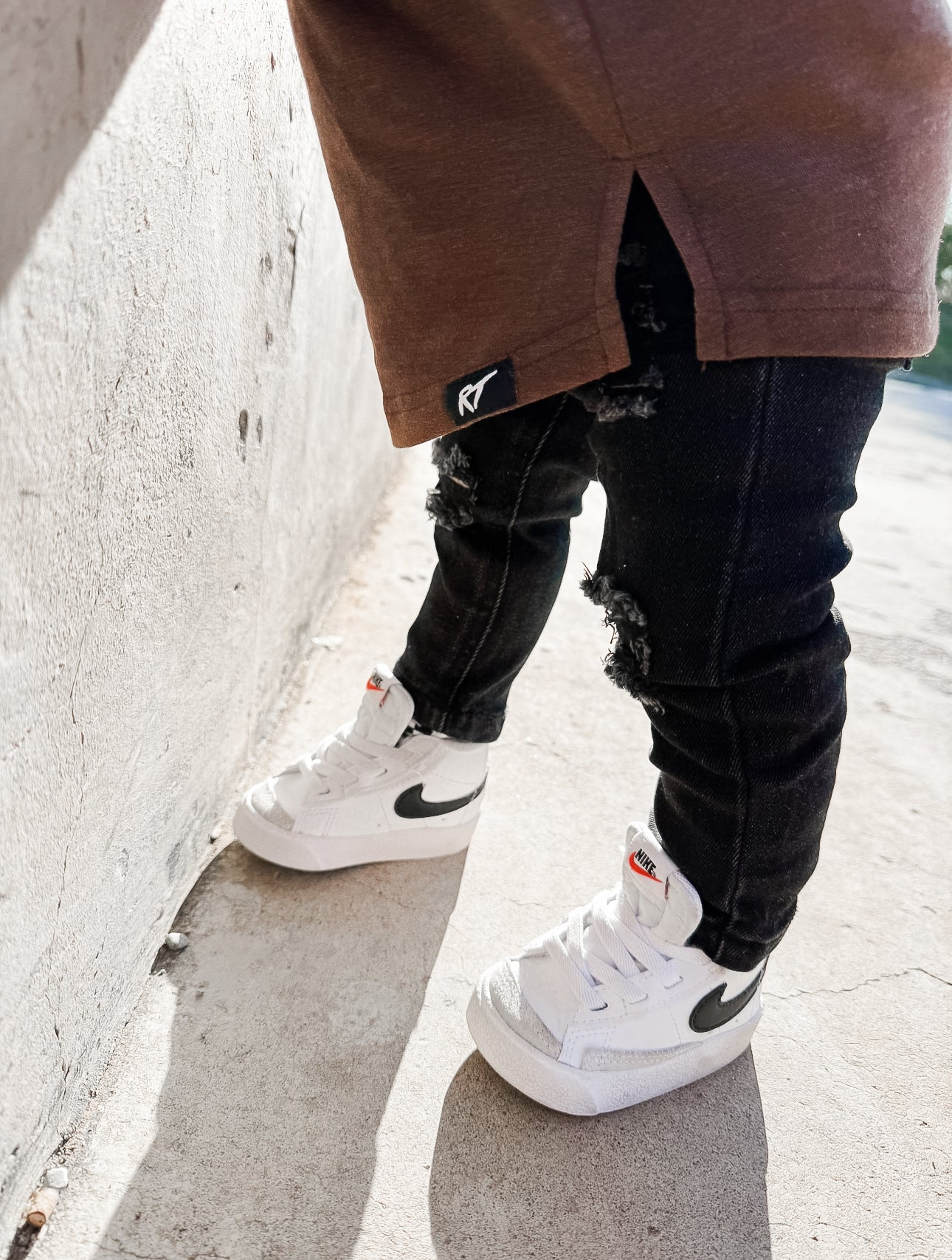 boy wearing a brown long sleeve tee, black ripped jeans, and white sneakers with a logo on a concrete surface.