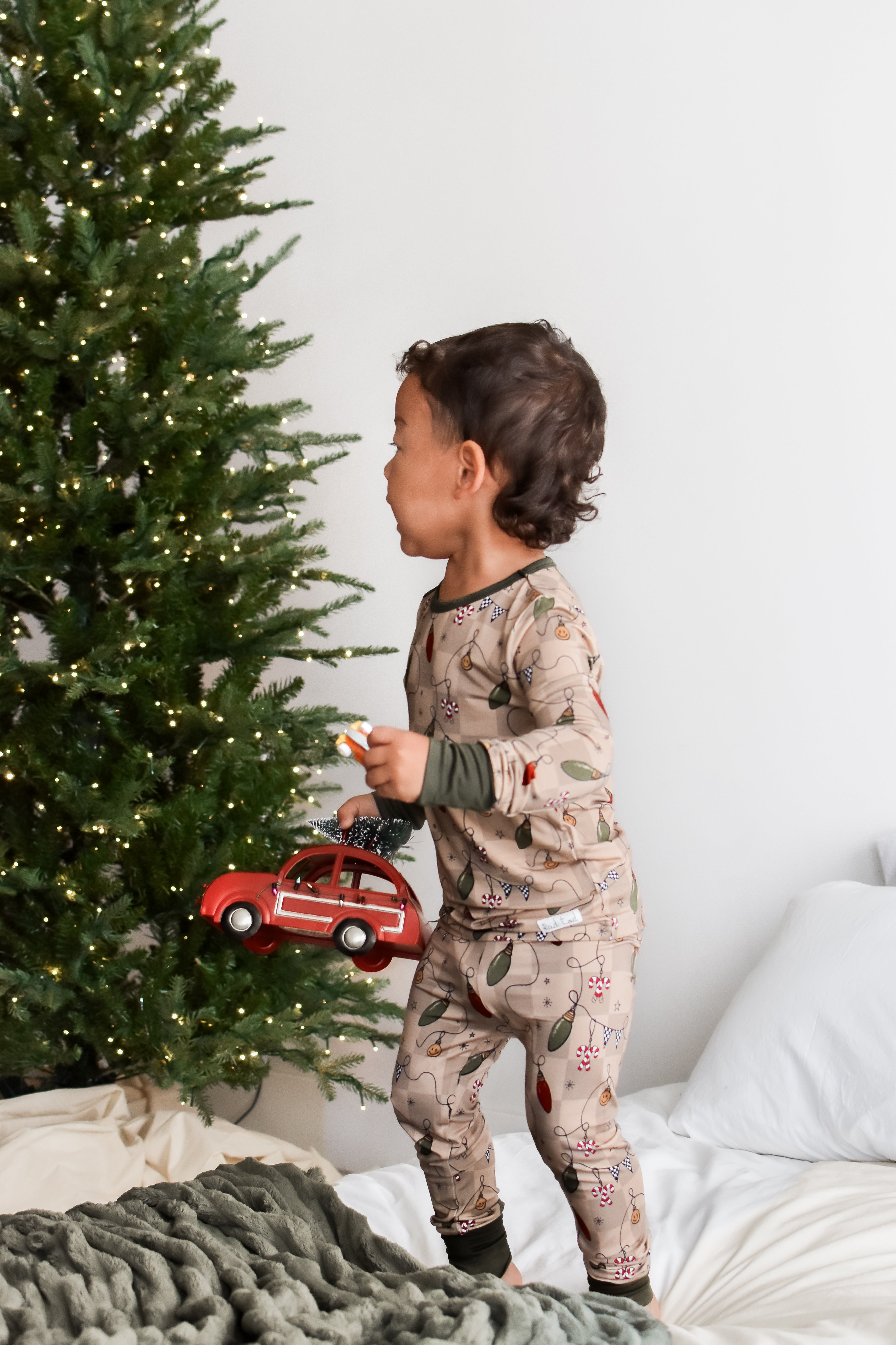 A toddler stands on a bed wearing a beige holiday-themed pajama set with a retro light print, holding a red toy car next to a decorated Christmas tree.